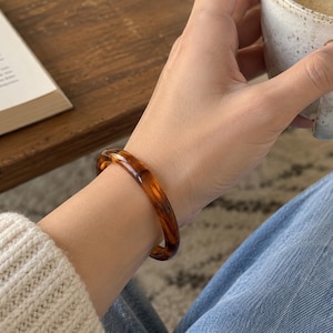 Puede incluir: Una pulsera rígida circular y lisa con un patrón translúcido moteado en tonos ámbar y marrón oscuro tipo carey. La pulsera tiene un acabado pulido y brillante con un perfil redondeado que rodea la muñeca con un grosor sólido y constante.