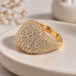 Puede incluir: Un anillo de sello de oro con una superficie superior ancha y redondeada, cubierta por completo con numerosas piedras pequeñas, claras y brillantes de corte redondo en pavé, con una banda de oro pulida y lisa.