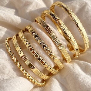 Puede incluir: Un conjunto de siete brazaletes y pulseras de metal en tono dorado. La colección incluye un diseño de cuerda trenzada, una banda con motivos de estrellas, un patrón geométrico, un diseño de eslabones en forma de corazón, una textura de rejilla y formas de media luna.