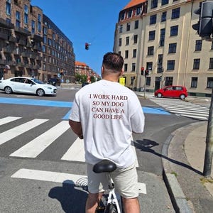 Könnte beinhalten: Ein weißes kurzärmeliges T-Shirt mit dunkelrotem Text auf der Rückseite, der lautet: I WORK HARD SO MY DOG CAN HAVE A GOOD LIFE. Das T-Shirt wird mit einer hellen Shorts kombiniert, während man auf einem Fahrrad durch eine Stadtstraße fährt.