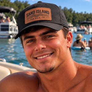 May include: A black baseball cap featuring a rectangular tan leather patch on the front. The patch is embossed with the text SAND ISLAND and TEN MILE, TN, accompanied by three wavy lines representing water, centered above and below the location name.