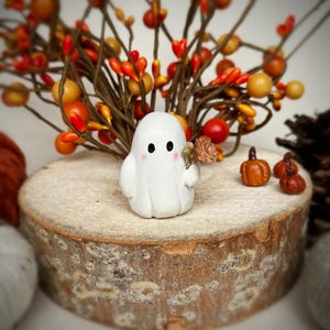 May include: A small white ghost figurine with black eyes and pink cheeks, holding a tiny brown flower. The ghost is positioned on a round wooden log slice, accompanied by three miniature orange pumpkins and decorative autumn branches.