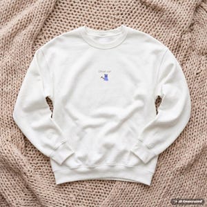 Peut inclure: Un sweat-shirt blanc à manches longues et col rond, orné d'un petit motif central représentant un chat fantôme violet clair. Au-dessus du chat, le texte Ghost cat est inscrit dans une police simple et sombre. Col, poignets et ourlet côtelés.