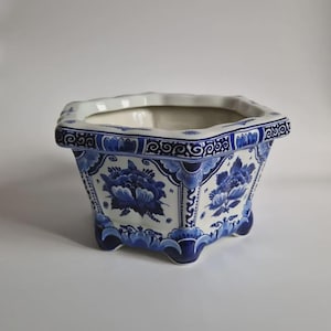 Könnte beinhalten: Ein quadratischer Keramikübertopf mit weißem Grund und komplexen blauen Blumenmustern sowie Schnörkelverzierungen. Das Gefäß hat einen gewellten Rand, eine weite Öffnung und steht auf vier kleinen, geschwungenen Füßen mit blauen Details.