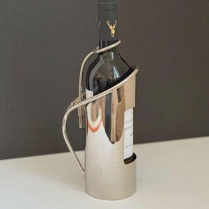 Pode incluir: Um suporte para garrafa de vinho em metal prateado polido com uma alça curva e um corpo cilíndrico que apresenta uma seção recortada. O suporte contém uma garrafa de vinho de vidro escuro com um rótulo preto com um emblema de cervo dourado.