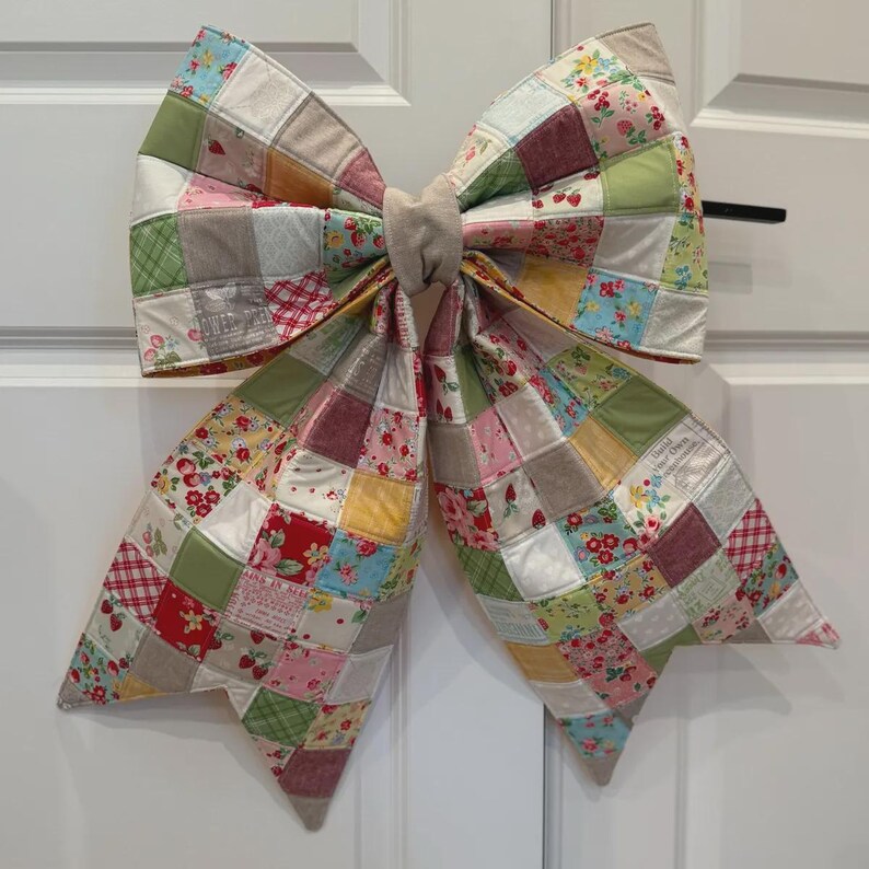 K&ouml;nnte beinhalten: Eine gro&szlig;e dekorative Schleife aus einem Patchwork aus gesteppten Stoffquadraten. Der Stoff weist eine Vielzahl von Mustern auf, darunter florale Drucke, Erdbeeren und Karomuster in den Farben Rot, Gr&uuml;n, Gelb, Rosa und Beige.