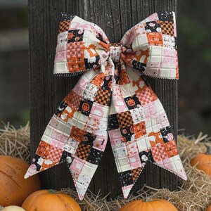K&ouml;nnte beinhalten: Eine gro&szlig;e dekorative Schleife aus gestepptem Stoff mit einem Patchwork-Muster aus orangefarbenen, schwarzen, wei&szlig;en und rosafarbenen Quadraten. Der Stoff zeigt Halloween-Motive wie Geister, K&uuml;rbisse, Spinnennetze und den Text trick or treat.