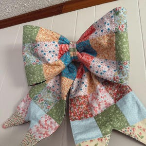 K&ouml;nnte beinhalten: Eine gro&szlig;e dekorative Schleife aus einem Patchwork aus gesteppten Stoffquadraten. Der Stoff zeigt eine Vielzahl von Blumen- und Pflanzenmustern in den Farben Hellblau, Salbeigr&uuml;n, sanftem Orange, Rot und Wei&szlig;, die in einem Raster angeordnet sind.