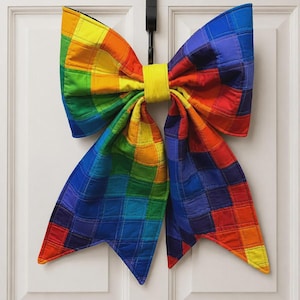 May include: A large, quilted fabric bow featuring a vibrant rainbow grid pattern of red, orange, yellow, green, blue, and purple squares. The bow is constructed with a central yellow fabric band and has two long, pointed tails hanging downwards.