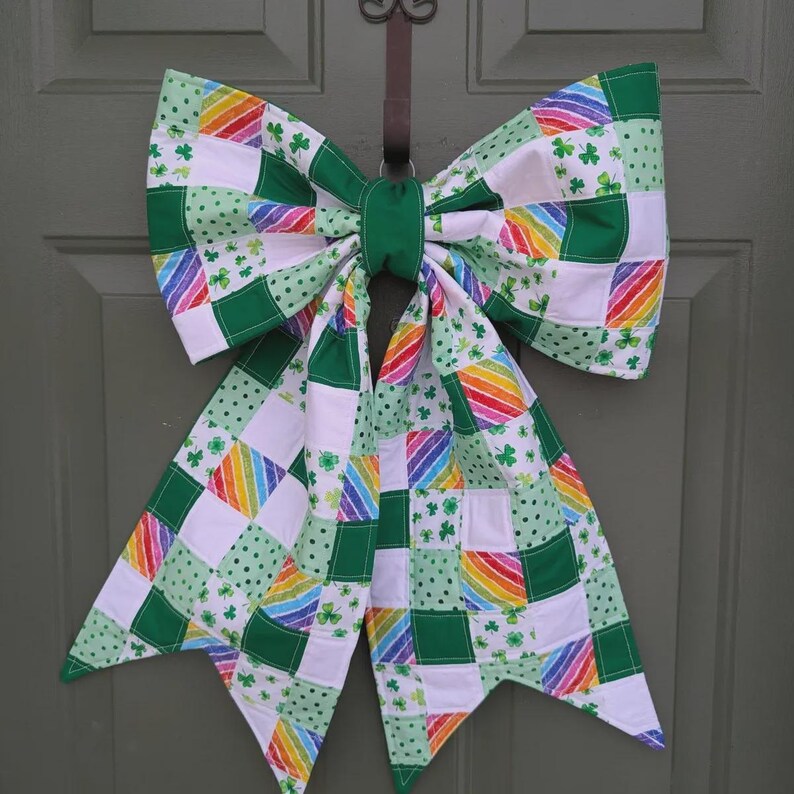 K&ouml;nnte beinhalten: Eine gro&szlig;e dekorative Schleife aus einem Patchwork aus Stoffquadraten. Der Stoff zeigt Muster aus gr&uuml;nen Kleebl&auml;ttern auf Wei&szlig;, einfarbigem Gr&uuml;n und diagonalen Regenbogenstreifen. Die Schleife hat zwei breite Schlaufen und zwei lange, spitze Enden.
