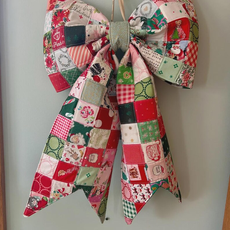 K&ouml;nnte beinhalten: Eine gro&szlig;e dekorative Schleife aus einem Patchwork aus gesteppten Stoffquadraten mit verschiedenen weihnachtlichen Motiven, darunter der Weihnachtsmann, Rentiere, Weihnachtssterne und Text mit Fresh Hot Cocoa und Home Sweet Holidays.