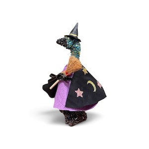 May include: A decorative goose figurine dressed as a witch, featuring a black pointed hat, a purple felt cape with a black overlay adorned with glittery stars and a crescent moon, and a small broomstick held under one wing, with a textured body.