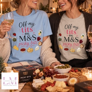 May include: A light blue t-shirt and a beige t-shirt, both featuring the black and pink text 'Off my tits on M&S picky bits' surrounded by illustrations of strawberries, grapes, olives, cheese, crackers, and a bowl of dip.