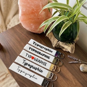 May include: Five white fabric keychains with metal rings, each featuring the word Typographie in different black fonts, including one with a red heart replacing the letter O. The hardware finishes vary in colour, including iridescent, bronze, gold, silver, and black.
