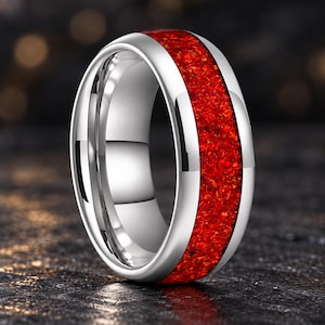 Könnte beinhalten: Ein polierter silberfarbener Ring mit einer breiten, mittigen Einlage aus zerkleinertem rotem Material mit texturierter, funkelnder Optik. Das Band hat ein glattes, abgerundetes Außenprofil und ein reflektierendes metallisches Finish.