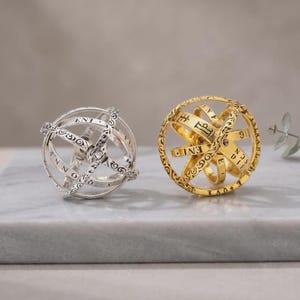 May include: Two spherical armillary rings, one in silver and one in gold, featuring intricate engraved scrollwork patterns and etched lettering along the bands. These adjustable metal spheres are designed to unfold into a ring shape.