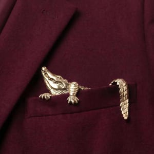 May include: A gold-toned metal alligator brooch attached to the breast pocket of a deep burgundy blazer. The alligator figure is positioned so its head and front legs emerge from the pocket, while its curved tail rests on the pocket edge.