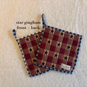 May include: Two square quilted pot holders featuring a red and brown gingham pattern with navy blue stars. Each pot holder has a navy blue and white checkered border and a small fabric loop. A white label with the text tcd+c is on one corner.