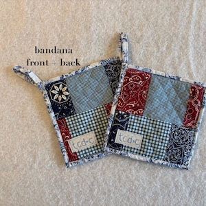May include: Two square quilted potholders featuring a patchwork design of blue, white, and red bandana-style patterns. Each potholder has a small fabric loop for hanging and a white rectangular label with the text tcd-c stitched on the front.