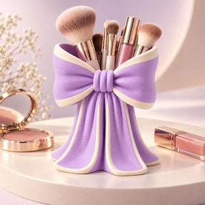 May include: A lavender and cream makeup brush holder shaped like a large bow with draped fabric details. The holder contains several makeup brushes with rose gold ferrules and brown bristles, alongside a rose gold lipstick tube.