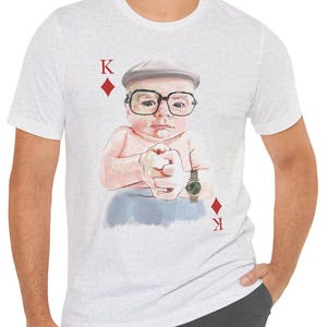 Può includere: Una t-shirt grigio melange chiaro con la grafica di un bambino che indossa un berretto piatto e occhiali con montatura spessa, in stile re di quadri. Il design include la lettera K e un simbolo di quadri rosso negli angoli superiore e inferiore.