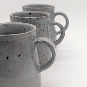 May include: Three ceramic mugs arranged in a diagonal line, each featuring a light gray glaze with dark brown speckles throughout. The mugs have a cylindrical body, a slightly flared rim, and a curved handle attached to the side of each piece.