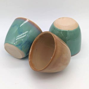May include: Three ceramic cups with a smooth, rounded shape. One cup is a glossy, variegated teal and blue, while the other two feature a warm, solid tan glaze. Each cup has an unglazed, light beige base and a simple, handleless design.
