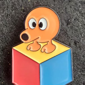 May include: A metal enamel pin featuring a small orange cartoon character with large white eyes sitting on top of a three-dimensional cube with red, yellow, and blue sides. A copper one-penny coin is placed above the pin for scale.