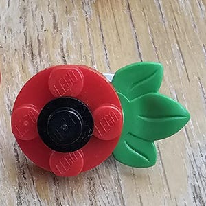 May include: Two lapel pins constructed from red and green plastic building blocks. One pin shows a black circular centre piece on a red base with green leaves, while the other displays the silver metal clutch backing and pin mechanism.