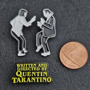 May include: Two enamel pins depicting figures in a dancing pose, one in a white shirt and black trousers, the other in a black suit. A third rectangular pin features the yellow text WRITTEN AND DIRECTED BY QUENTIN TARANTINO. A one-penny coin is shown for scale.