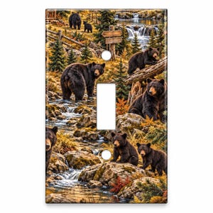 May include: A rectangular light switch cover plate featuring a detailed forest scene with multiple black bears near a flowing stream, waterfalls, evergreen trees, and a wooden signpost. The design includes rocky terrain and autumn-toned foliage.