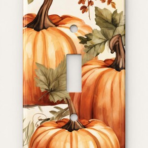 May include: A rectangular light switch cover plate featuring a watercolor-style illustration of orange pumpkins with brown stems and green leaves against a cream background. The plate has a central rectangular cutout and two circular screw holes.