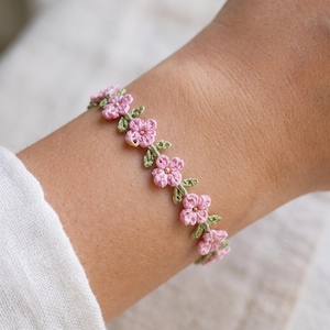 May include: A bracelet featuring a series of small, crocheted pink flowers with green leaves. Each flower has a tiny, metallic gold-colored bead at its center. The floral chain is constructed from fine thread and wraps around the wrist.