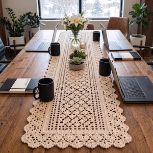 May include: A long, rectangular, cream-coloured crochet table runner with an intricate openwork pattern and a scalloped edge, centred on a wooden table alongside black mugs, laptops, notebooks, a glass vase with white lilies, and succulents.