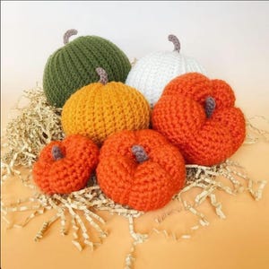 May include: A collection of six crocheted pumpkins in various sizes and colors, including olive green, white, yellow-orange, and bright orange. Each pumpkin features a small, textured brown stem at the top and a distinct ribbed or bumpy surface.