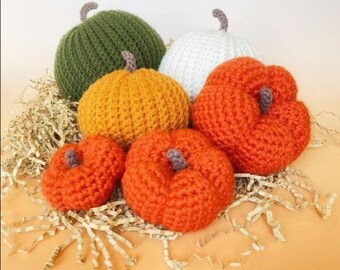 May include: A collection of six crocheted pumpkins in various sizes and colors, including olive green, white, yellow-orange, and bright orange. Each pumpkin features a small, textured brown stem at the top and a distinct ribbed or bumpy surface.