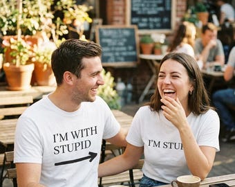 May include: Two white t-shirts featuring black text. One shirt displays the text I'M WITH STUPID with a black arrow pointing to the right. The other shirt displays the text I'M STUPID. Both shirts have short sleeves and a crew neckline.