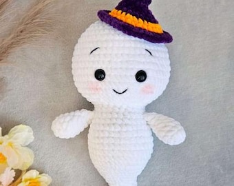 May include: A white crocheted ghost plush toy featuring a purple witch hat with an orange band, black plastic safety eyes, a small embroidered mouth, and soft pink blush on the cheeks. The ghost has a rounded head and a tapered body shape.