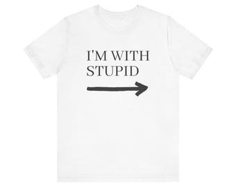 May include: A white short-sleeved t-shirt featuring the black text I'M WITH STUPID printed on the chest, positioned above a black hand-drawn style arrow pointing toward the right side of the garment. The shirt has a crew neckline.