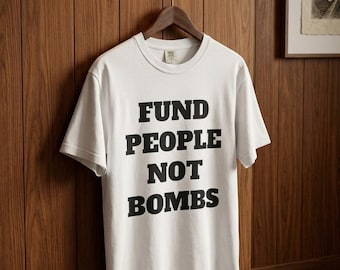 May include: A white short-sleeved t-shirt featuring the black text "FUND PEOPLE NOT BOMBS" printed in a bold, block-style font on the front. The garment is displayed on a wooden hanger against a vertical wood-paneled wall.