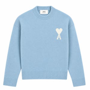 Puede incluir: Un suéter de manga larga en color azul claro con cuello redondo y acabados acanalados en el cuello, los puños y el dobladillo. El pecho presenta un logotipo bordado en blanco que consiste en un corazón sobre la letra A. La etiqueta interior dice ami.