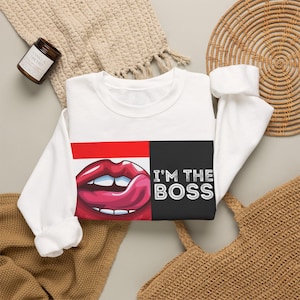 Könnte beinhalten: Ein weißes Langarm-Sweatshirt mit einem Grafikdruck von roten Lippen, bei dem eine Zunge über die Oberlippe leckt. Rechts neben den Lippen ist der Text "I'M THE BOSS" in weißen Großbuchstaben auf einem schwarzen rechteckigen Hintergrund gedruckt.