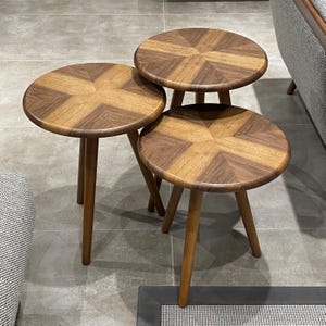 Peut inclure: Un ensemble de trois tables d'appoint rondes en bois avec un motif de placage en étoile sur les plateaux. Chaque table possède trois pieds fuselés et une finition lisse et polie, disposées en groupe pour montrer différentes hauteurs.