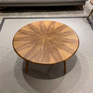 May include: A round wooden coffee table featuring a sunburst veneer pattern on the tabletop. The surface displays alternating light and dark brown wood grain segments radiating from the center. The table is supported by three tapered wooden legs.