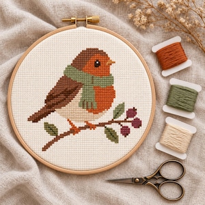 May include: A cross-stitch embroidery project in a round wooden hoop featuring a brown and orange bird wearing a green scarf, perched on a branch with purple berries. Three spools of embroidery floss and metal scissors are included.