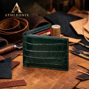 May include: A dark green bifold wallet with a textured crocodile skin pattern. The wallet is partially open, revealing red and black interior card slots. The brand name ATHLEONIX is printed in gold above the wallet with a stylized A logo.