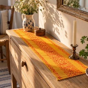 Pode incluir: Um caminho de mesa retangular num tom amarelo vibrante com uma faixa central de padrões geométricos e florais laranja complexos. O tecido tem uma trama texturizada com uma borda decorativa de franjas, estendido sobre uma superfície.
