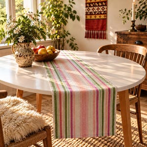 May include: A rectangular table runner featuring a vertical striped pattern in shades of pink, green, and cream. The fabric has a textured weave and hangs over the edge of a round white table, displaying a consistent multi-colored linear design.