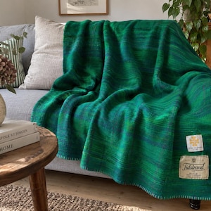 May include: A woven throw blanket in shades of emerald and forest green with subtle blue undertones. The blanket features a stitched edge and two rectangular fabric labels in the corner, one with a gold cross and one reading Tidstrand.