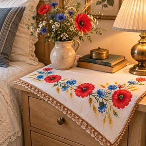 May include: A rectangular off-white table runner featuring a vibrant floral print of red poppies, blue cornflowers, and golden wheat stalks. The runner is finished with a delicate scalloped brown lace trim along the edges, adding texture to the fabric.